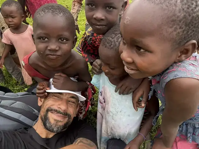 Leon lying on the grass surrounded by laughing children leaning into the camera
