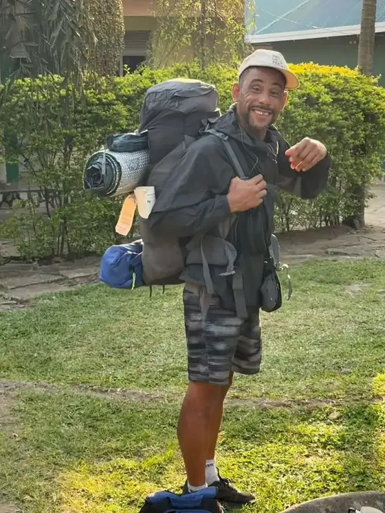 Leon standing side on with a full expedition backpack and bedroll, pointing at the camera