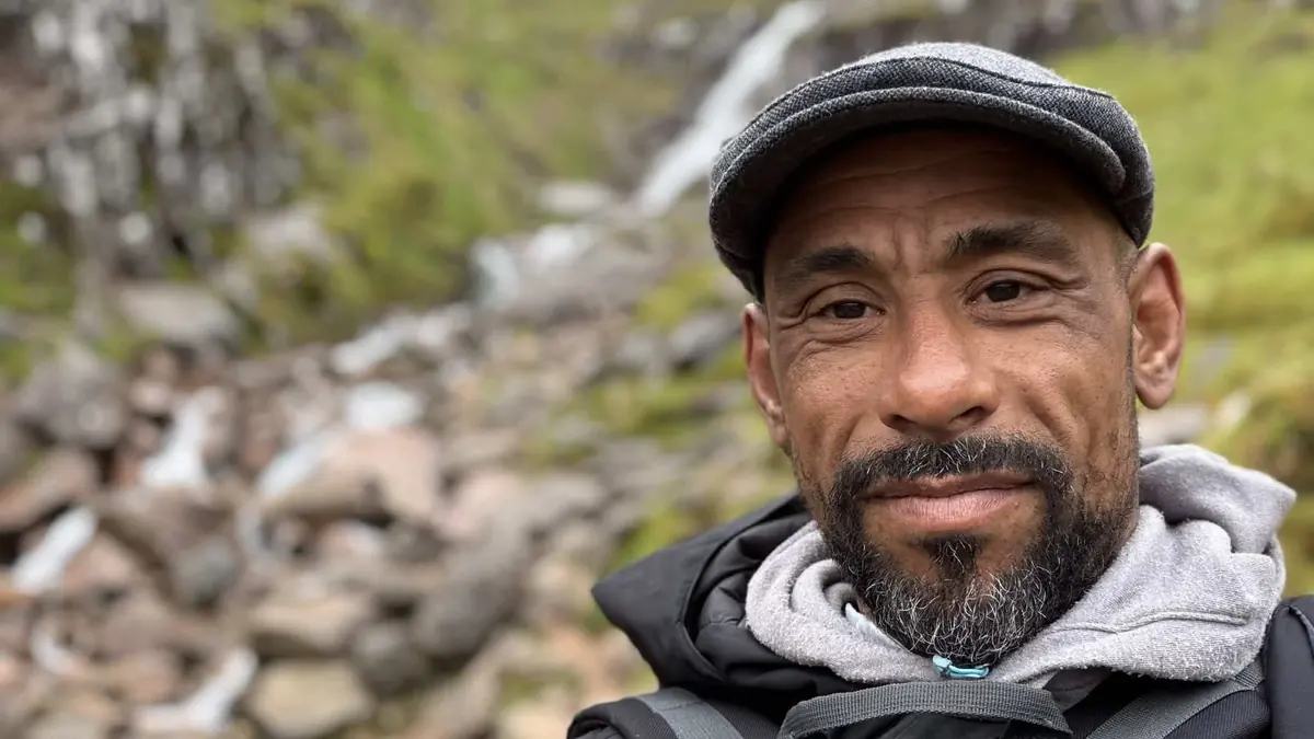 Portrait of Leon McFarlane in a flat cap, a waterfall and hillside behind him